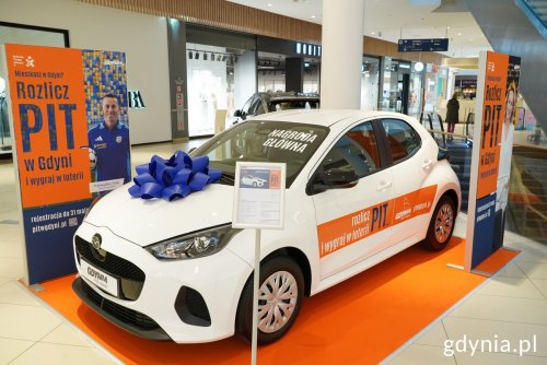 Wnętrze Centrum Riviera, pomarańczowy dywan, główny hol, stoi biały samochód Mazda 2 Hybrid z kokardą, obok ekspozytory z Rozlicz PIT w Gdyni