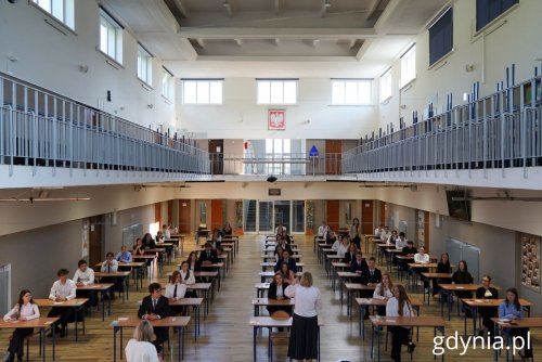 Aula w X Liceum Ogólnokształcącym w Gdyni, stoliki, przy nich kilkudziesięciu maturzystów, na stołach kartki z egzaminami