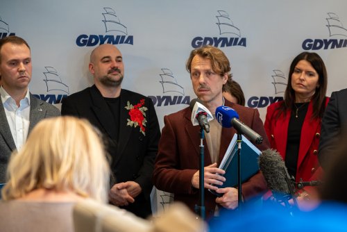Sala w Urzędzie Miasta Gdyni, grupa radnych, na środku do mikrofonów przemawia Łukasz Piesiewicz, w tle logotypy Gdyni