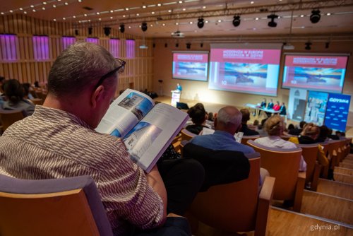 Sala konferencyjna w PPNT, widownia pełna ludzi, prelegenci na scenie, wyświetlane zdjęcia. Na pierwszym planie mężczyzna siedzący w ostatnim rzędzie przegląda książkę z projektami architektury