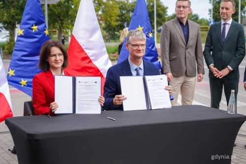 Przestrzeń na zewnątrz, czarny stół, przy stole Joanna Lech i Rafał Geremek prezentują podpisane umowy w teczkach, z tyłu kilka osób i flagi państwowe
