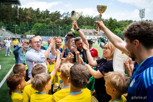 Murawa stadionu rugby w Gdyni, grupa dzieci w sportowych koszulkach, Oktawia Gorzeńska i Marcin Skwierawski wspólnie z dziećmi podnoszą puchar