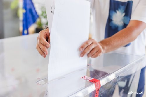 Lokal wyborczy, przezroczysta urna, męskie ręce wsuwają do urny kartę do głosowania