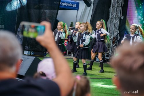 Scena w muszli koncertowej, widoczna publiczność, na scenie grupa dziewczynek w jednakowych strojach tańczy i śpiewa