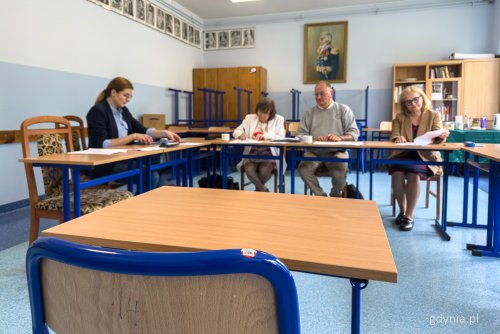 Sala lekcyjna w szkole, na środku sali ustawione stoliki, kilka osób siedzi w charakterze egzaminatorów, na stołach dokumenty