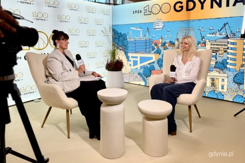 Studio filmowe, dwie kobiety siedzące w fotelach przemawiają do mikrofonów, w tle logotypy 100-lecia Gdyni i ścianka z grafiką 100-lecia