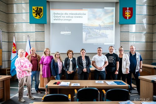 Sala obrad Rady Miasta Gdyni, grupa osób pozuje do zdjęcia z prezydent Gdyni Aleksandrą Kosiorek, w tle slajd z konferencji prasowej