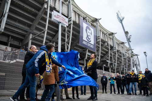 Stadion, tabliczka z nazwą placu, w tle zdjęcie Janusza Kupcewicza, grupa ludzi ściąga materiał i odsłania tablicę