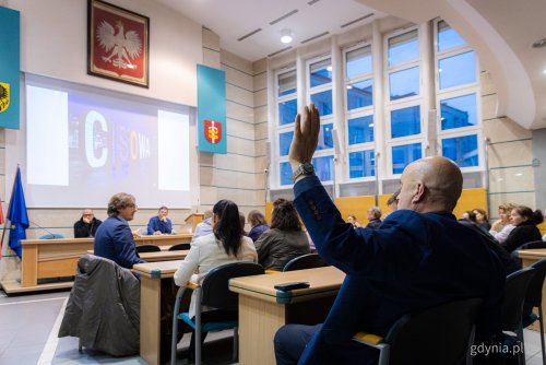 Sala obrad Rady Miasta Gdyni, ludzie w ławach, na ekranie wyświetlane zdjęcie, mężczyzna unosi rękę w górę.