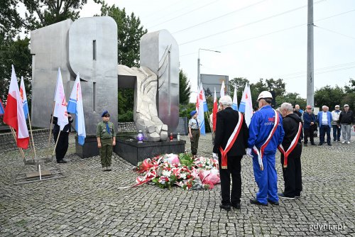 Pomnik Ofiar Grudnia 1970, plac, flagi Polski i Gdyni, dwie osoby składają kwiaty pod pomnikiem