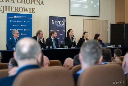 Sala audytoryjna w Wejherowie, na scenie stół prezydialny, siedzi kilka osób, wojewoda pomorskie przemawia przez mikrofon