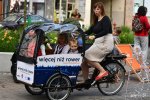 Kobieta prowadzi rower cargo, w środku skrzyni siedzą dzieci, w tle ulica