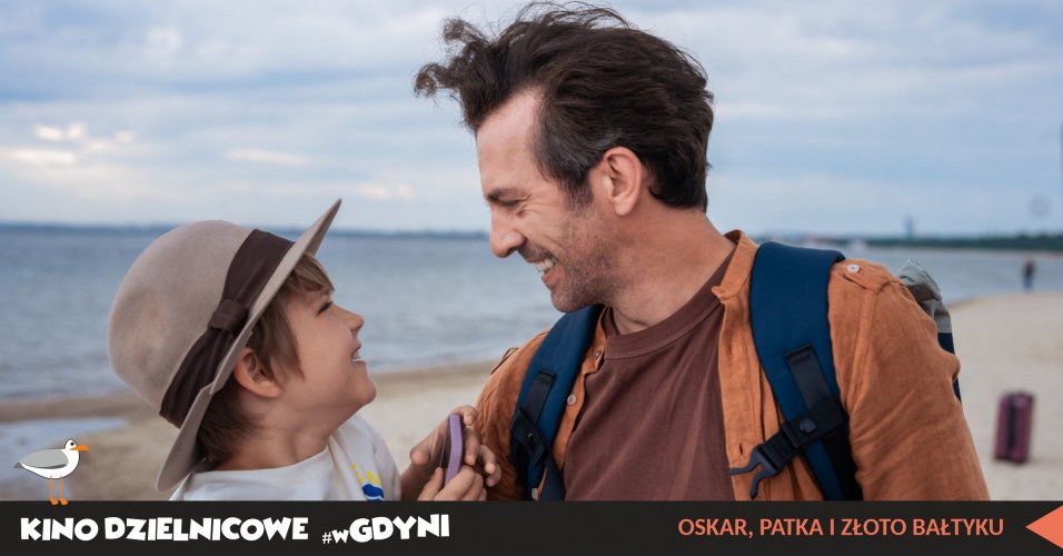 Kadr z filmu  „Oskar, Patka i złoto Bałtyku”. Mężczyzna c chłopcem w kapeluszy, uśmiechnięci na tle morza.