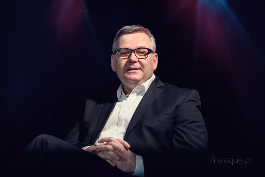 Mężczyzna siedzący w czarnej marynarce i białej koszuli, na czarnym tle. Spogląda w kierunku obiektywu i ma założone dłonie jedna na drugą.