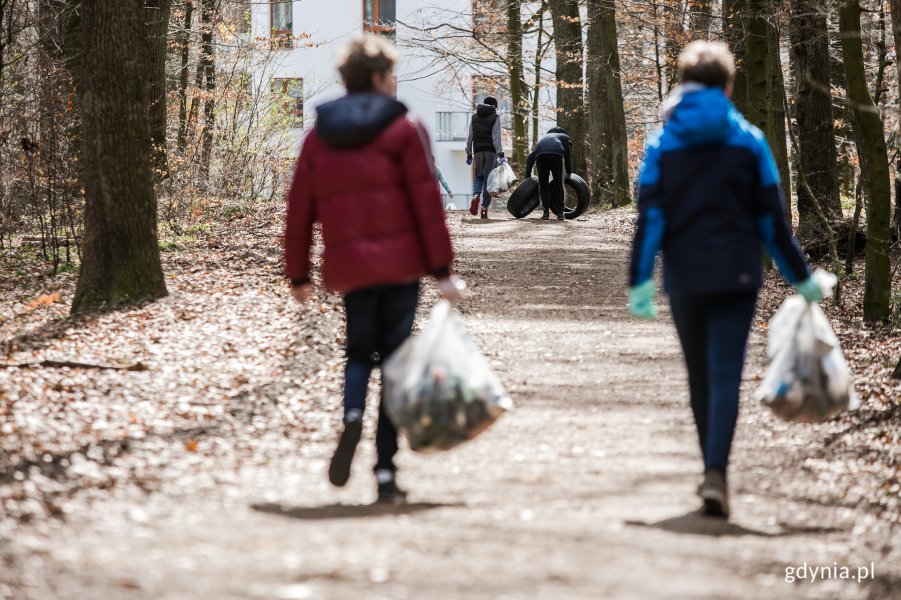 Dwie postacie, niosą worki pełne śmieci, idą przez las.