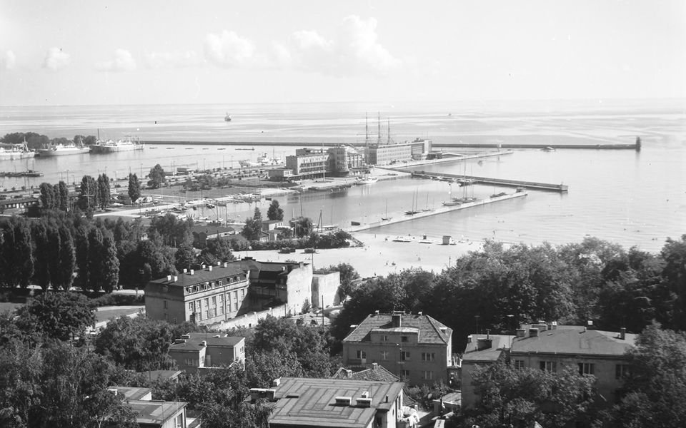 Czarno-białe zdjęcie Gdyni. Najprawdopodobniej wykonane w okolicach Kamiennej Góry. Na zdjęciu rozciąga się widok na morze, plażę. Widać Akwarium Gdyńskie oraz koniec Mola Południowego.