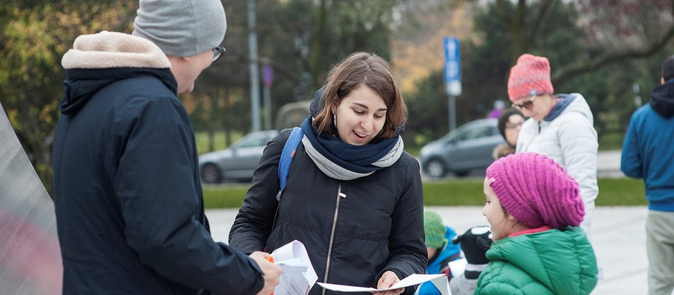 Uśmiechnięta kobieta rozmawiająca z dwójką ludzi na ulicy, trzyma w ręku ulotkę lub mapę.