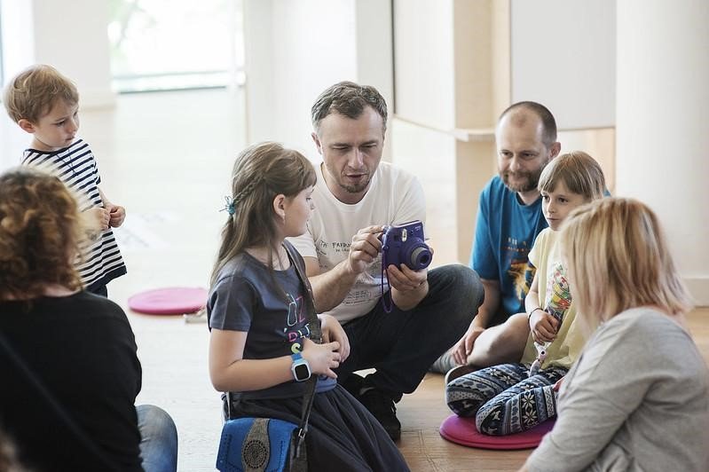 Mężczyzna trzymający w ręce aparat i tłumaczący coś zebranym dzieciom.
