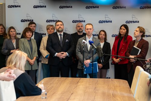 Sala w urzędzie miasta Gdyni, w tle logotypu miasta na białym tle, grupa radnych przemawia do mikrofonu, stół, fragment kamery