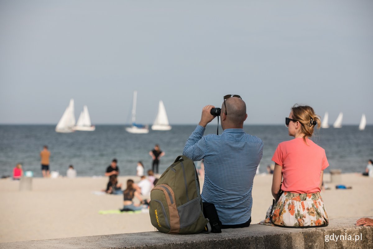 Plaża w Gdyni. Kobieta i mężczyzna siedzą, patrzą na przepływające w oddali żaglowce. Mężczyzna patrzy przez lornetkę. Widoczna plaża i morze