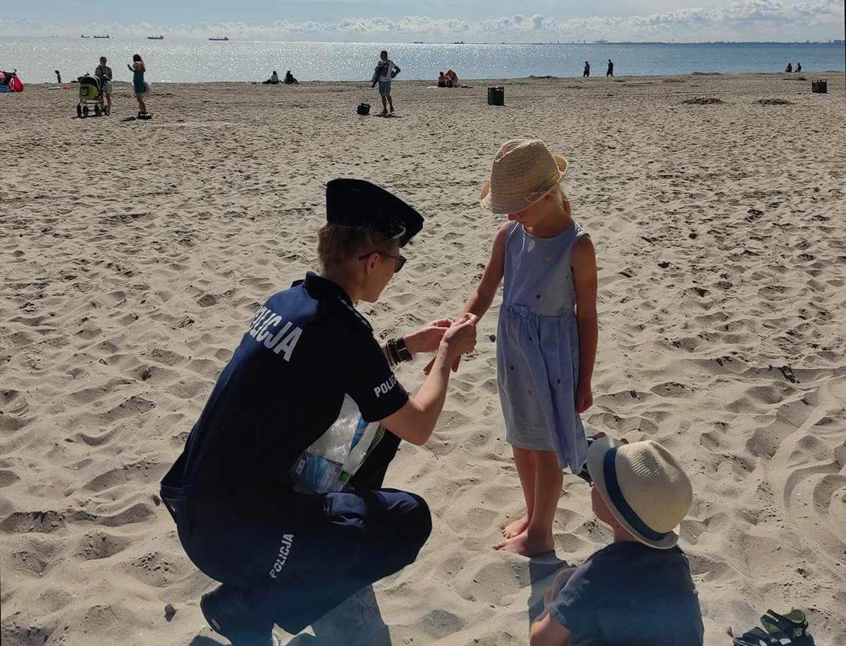 na zdjęciu policjantka kuca na plaży przy małej dziewczynce i zakłada jej niebieska opaskę. Przygląda się temu mały chłopiec. W tle morze i plażowicze
