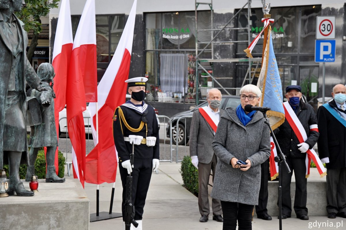 Na zdjęciu przemawia Joanna Zielińska, przewodnicząca Rady Miasta Gdyni. W drugim planie mundurowi Marynarki Wojennej. Trzymają proporce z flagami biało-czerwonymi.