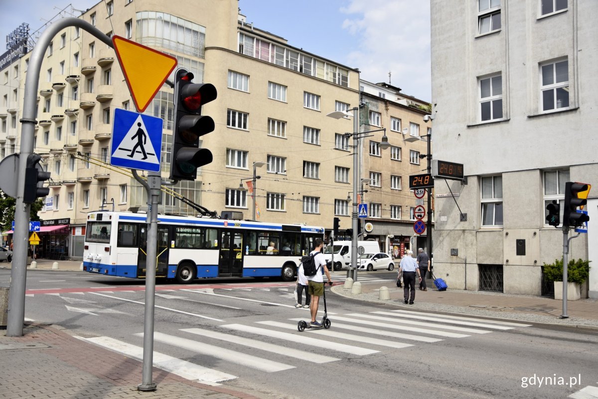 Na zdjęciu widoczne skrzyżowanie alei Piłsudskiego z ulicą Świętojańską w Gdyni i róg budynku Urzędu Miasta Gdyni. Przez przejście dla pieszych przejeżdża hulajnoga, w tle ulicą Świętojańską jedzie trolejbus