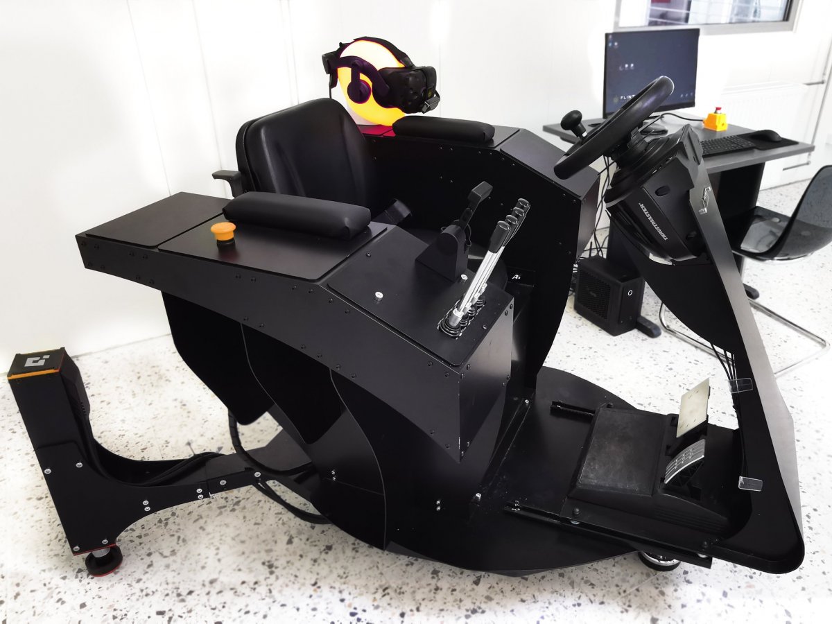 Czarny fotel - symulator VR z googlami i dżojstikami do sterowania, w tle białe pomieszczenie, biurka