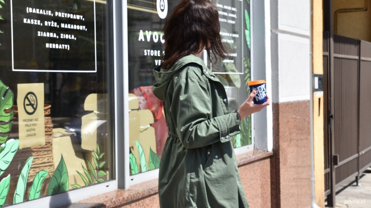 Kobieta trzyma w prawej ręce kubek miejski z napisem: Gdynia. Stoi tyłem na tle witryny sklepu Avocado przy ul. Starowiejskiej. Fot. Magdalena Czernek