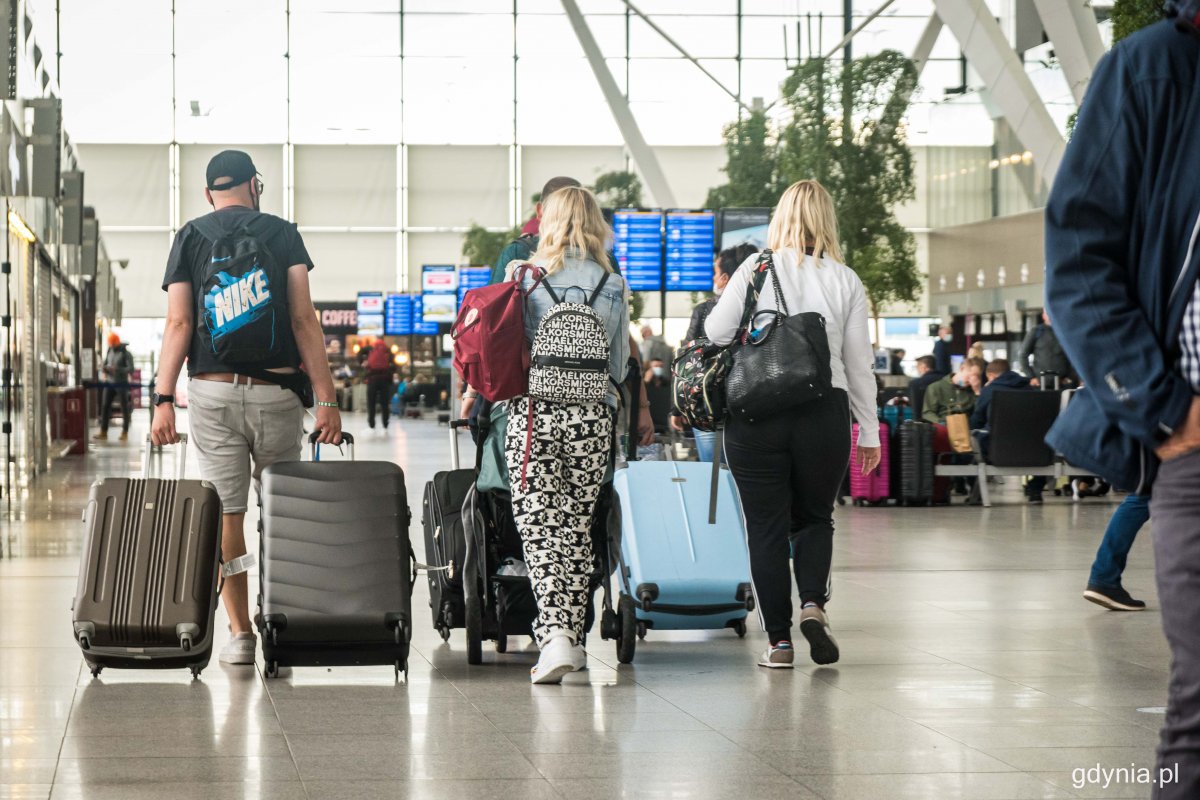 Terminal pasażerki w Porcie Lotniczym Gdańsk. Troje dorosłych ludzi idzie przez terminal, ciągną walizki lotnicze. W tle tablica z odlotami, ławki, sklepy i pasażerowie