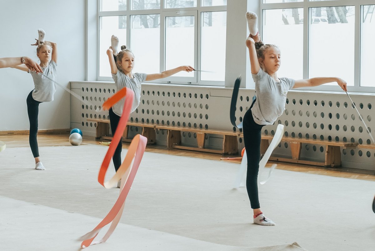 Sala gimnastyczna, w tle wielkie okna, na podłodze białe maty, na zdjęciu dziewczynki ćwiczą układ gimnastyczny z szarfami, stojąc z jedną nogą w górze.