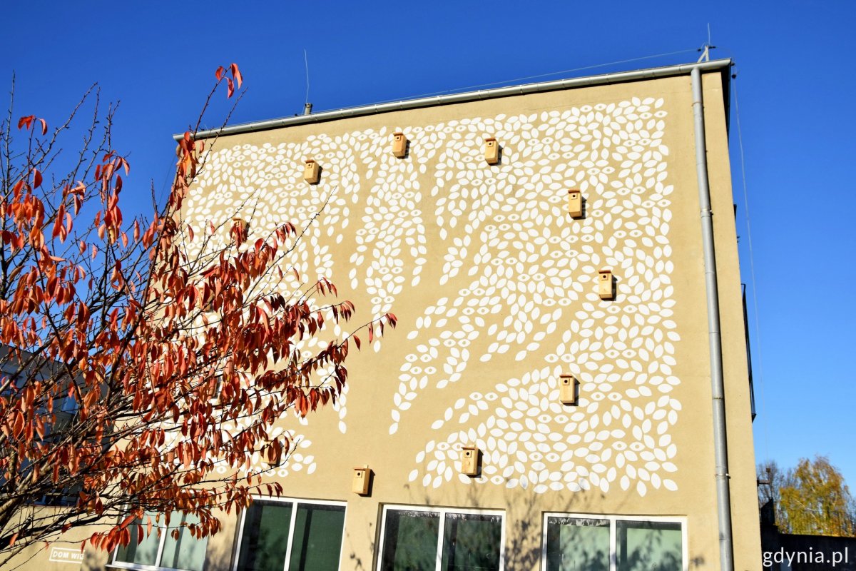 Ściana budynku SP nr 23 w Gdyni z muralem w kształcie drzewa, na ścianie budki dla ptaków, widoczne drzewo z jesiennymi liściami