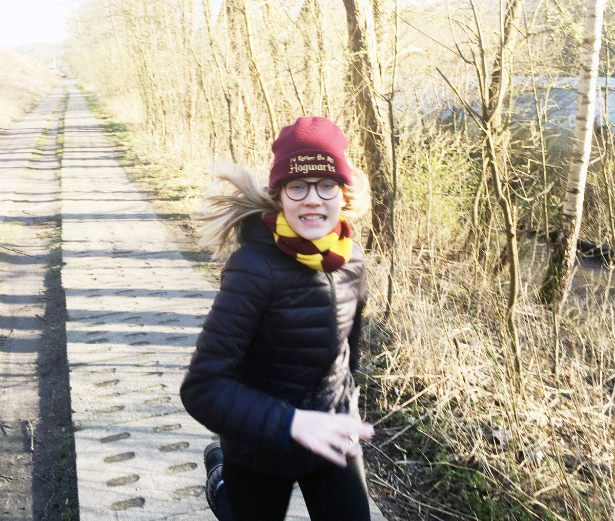 Dziewczynka z jasnymi włosami, w czapce i kurtce, biegnie przed siebie.