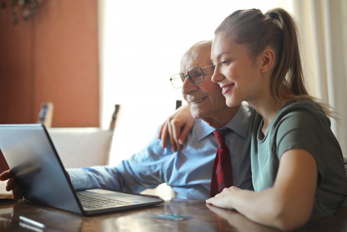 Senior i młoda kobieta siedzą razem przed laptopem. Oboje się uśmiechają. Kobieta obejmuje mężczyznę ramieniem
