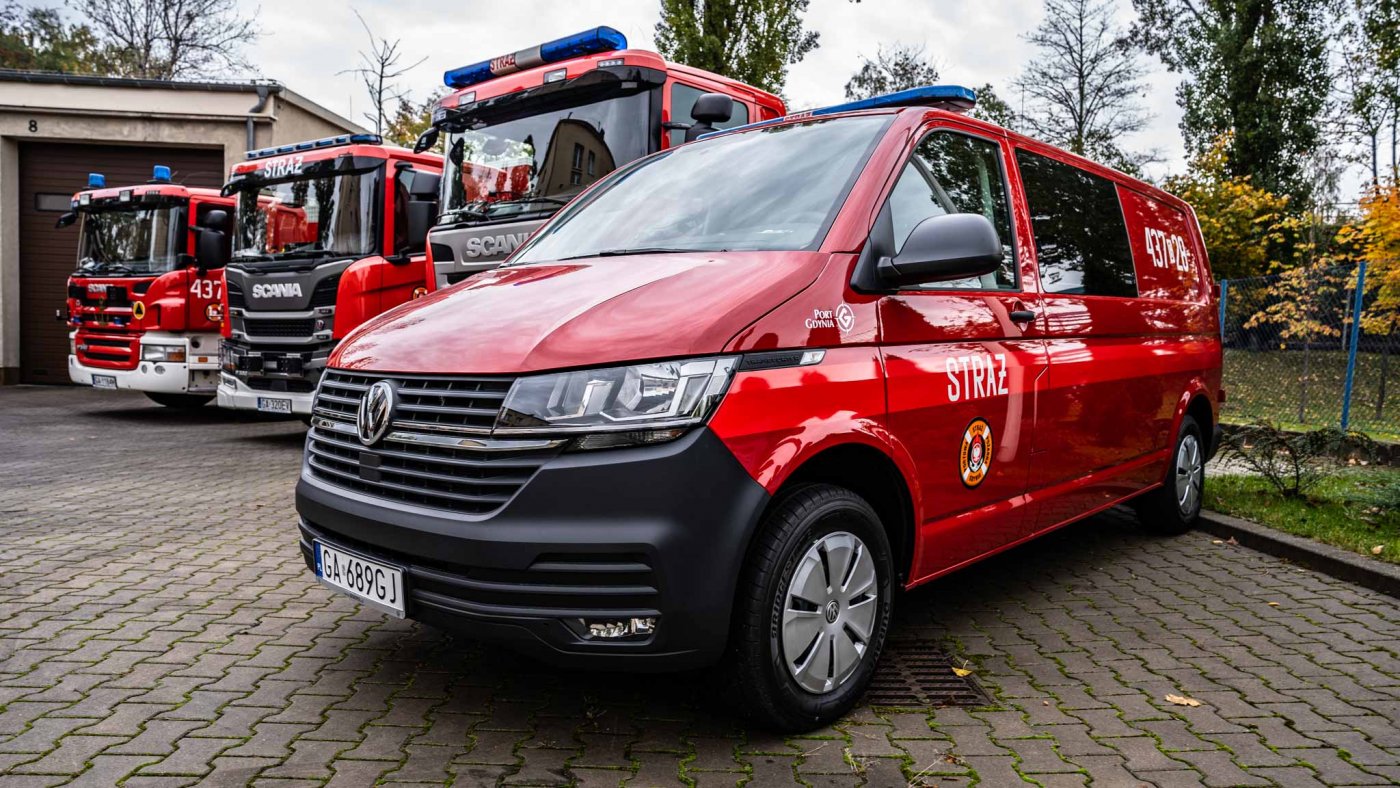 Na zdjęciu zaparkowany na placu nowy wóz typu bus dla straży pożarnej, w czerwonym kolorze, z symbolami Portowej Straży Pożarnej. Obok widoczny fragment wozu bojowego