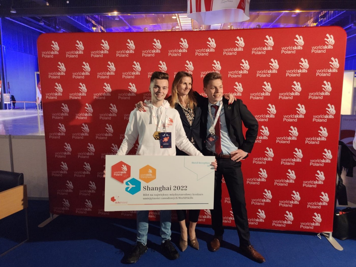Dwóch młodych finalistów, jeden z nich trzyma duże zaproszenie do Szanghaju, w towarzystwie nauczycielki.W tle ścianka z logo zawodów. Fot. ZSHG.