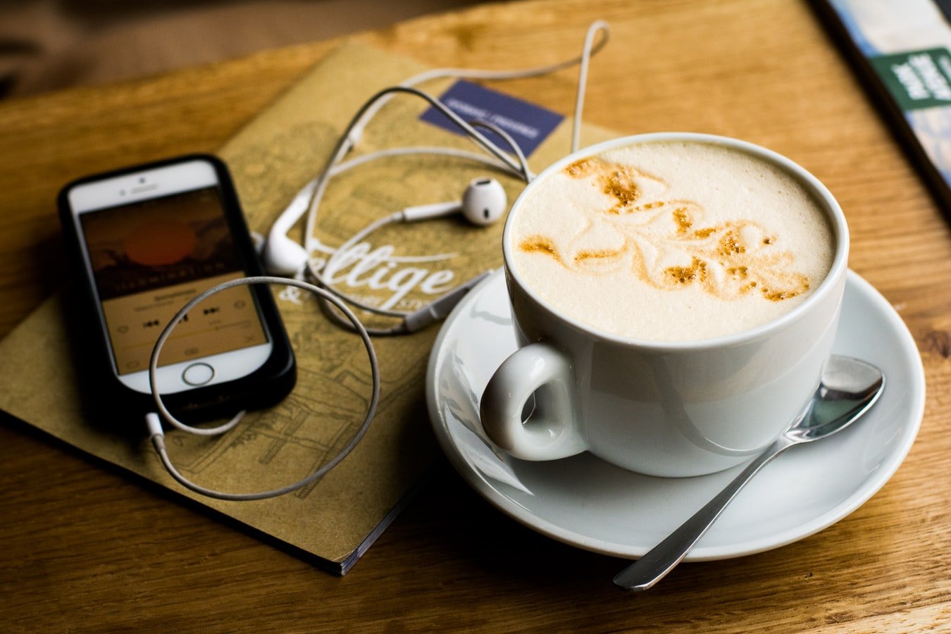 Na zdj. filiżanka z kawą, obok telefon komórkowy ze słuchawkami i notatnik. Zdj. ilustracyjne ze strony unsplash.com.