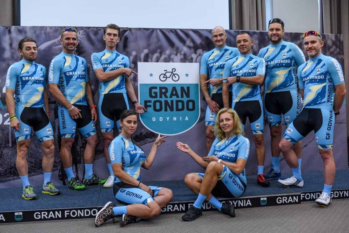 Zawodnicy (kobiety i mężczyźni) w błękitnych strojach stojący w dwóch szeregach przy znaku imprezy Gran Fondo Gdynia 2021.