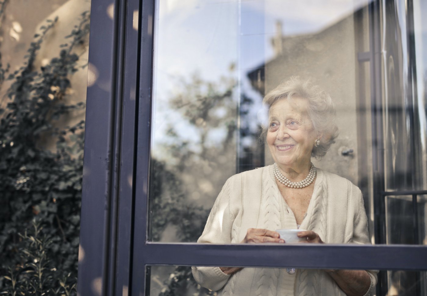 Starsza, uśmiechnięta kobieta widziana zza szyby okna swojego domu. Ma siwe włosy, ubrana jest w jasny, kremowy sweter, w dłoniach trzyma białą filiżankę z porcelany.