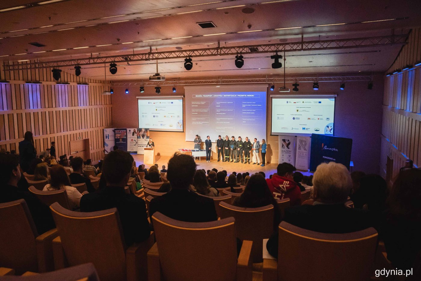 Sala konferencyjna w PPNT Gdynia. Na scenie w rzędzie pozuje do zdjęcia grupa młodzieży. Na widowni ludzie. Na ekranach wyświetla się nazwa konkursu 