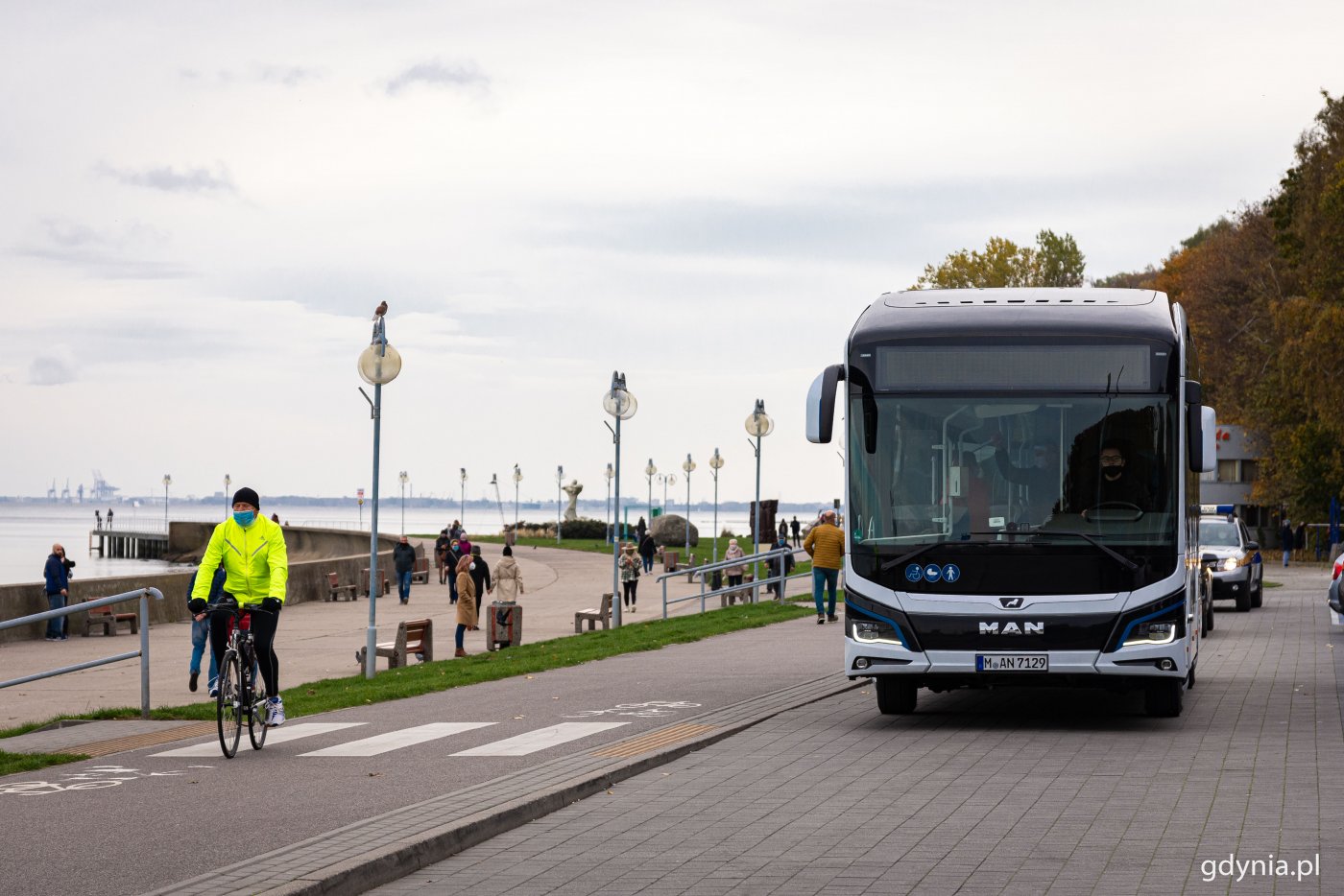Elektryczny autobus na parkingu przy Skwerze Arki Gdyni, obok na drodze rowerowej przygląda mu się rowerzysta, spacerują ludzie. W tle widoczny bulwar nadmorski i morze.