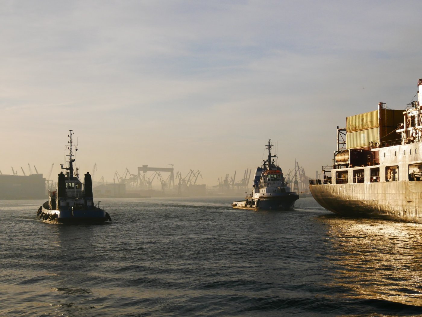 Basen portowy Portu Gdynia. Widoczne nabrzeża, na wodzie dwa holowniki i część statku handlowego kontenerami na pokładzie
