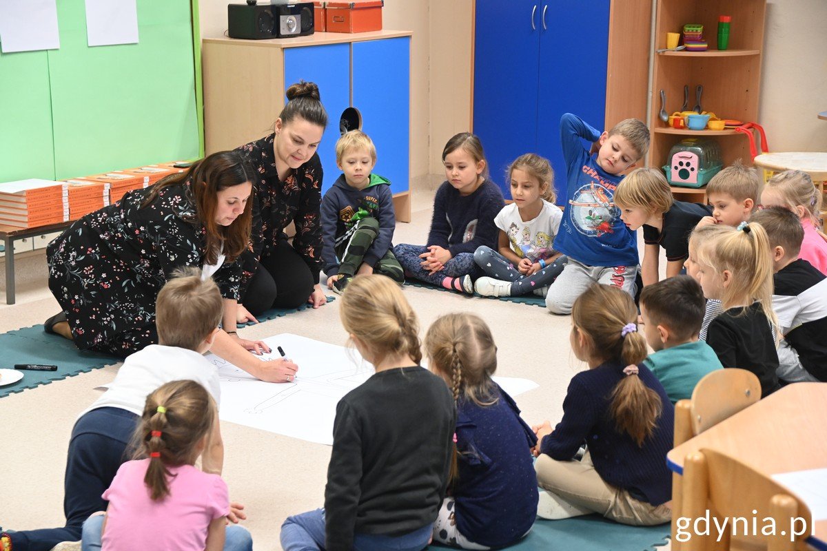 Na zdjęciu znajduje się grupa przedszkolaków ubranych różnokolorowo i dwie panie nauczycielki w ciemnych ubraniach. Grup dzieci siedzi na piankowych puzzlach i zajęta jet rysowaniem człowieka na dużej kartce papieru. Dzieci znajdują się w kolorowej sali Przedszkola nr 48 w Gdyni