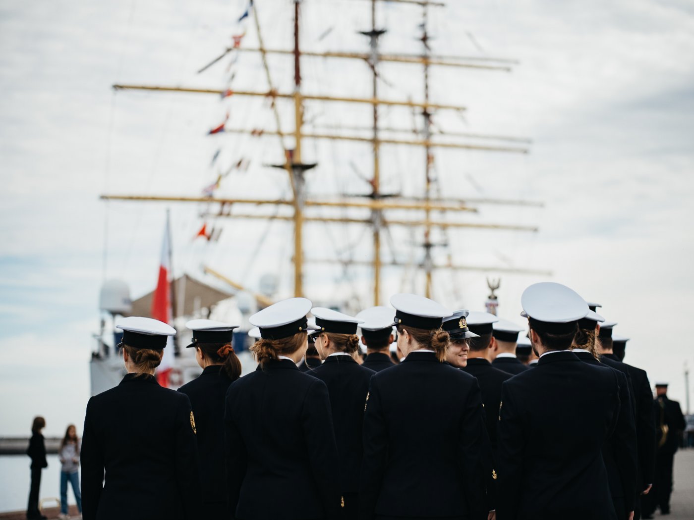 Nabrzeże Pomorskie w Gdyni. Stoi odwrócona plecami grupa studentów Uniwersytetu Morskiego w granatowych mundurach i białych czapkach marynarskich, za nimi widoczne żagle statku