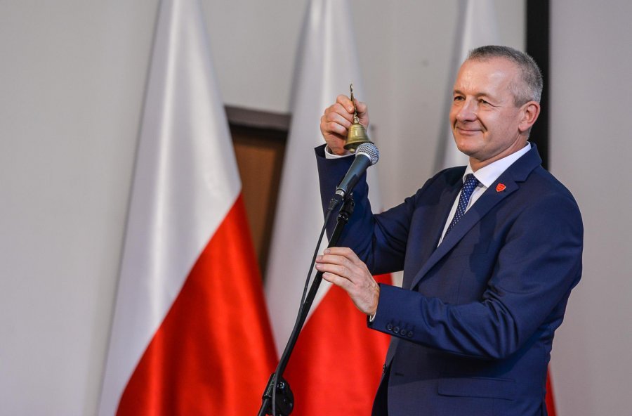 Na zdjęciu w tle flagi polski, na pierwszym planie dyrektor Kosakowski z dzwonkiem, w niebieskim garniturze.