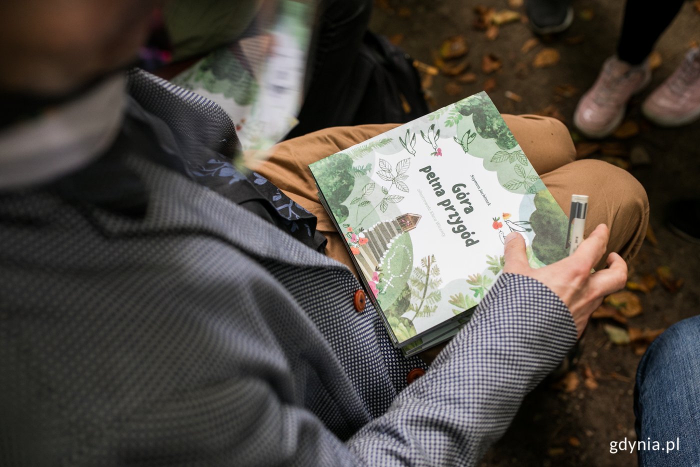 Ubrana w szarą marynarkę osoba widziana z góry. Nie widzimy jej twarzy ani głowy. Trzyma w rękach, na kolanach egzemplarz najnowszej Książeczki Gdynieczki.