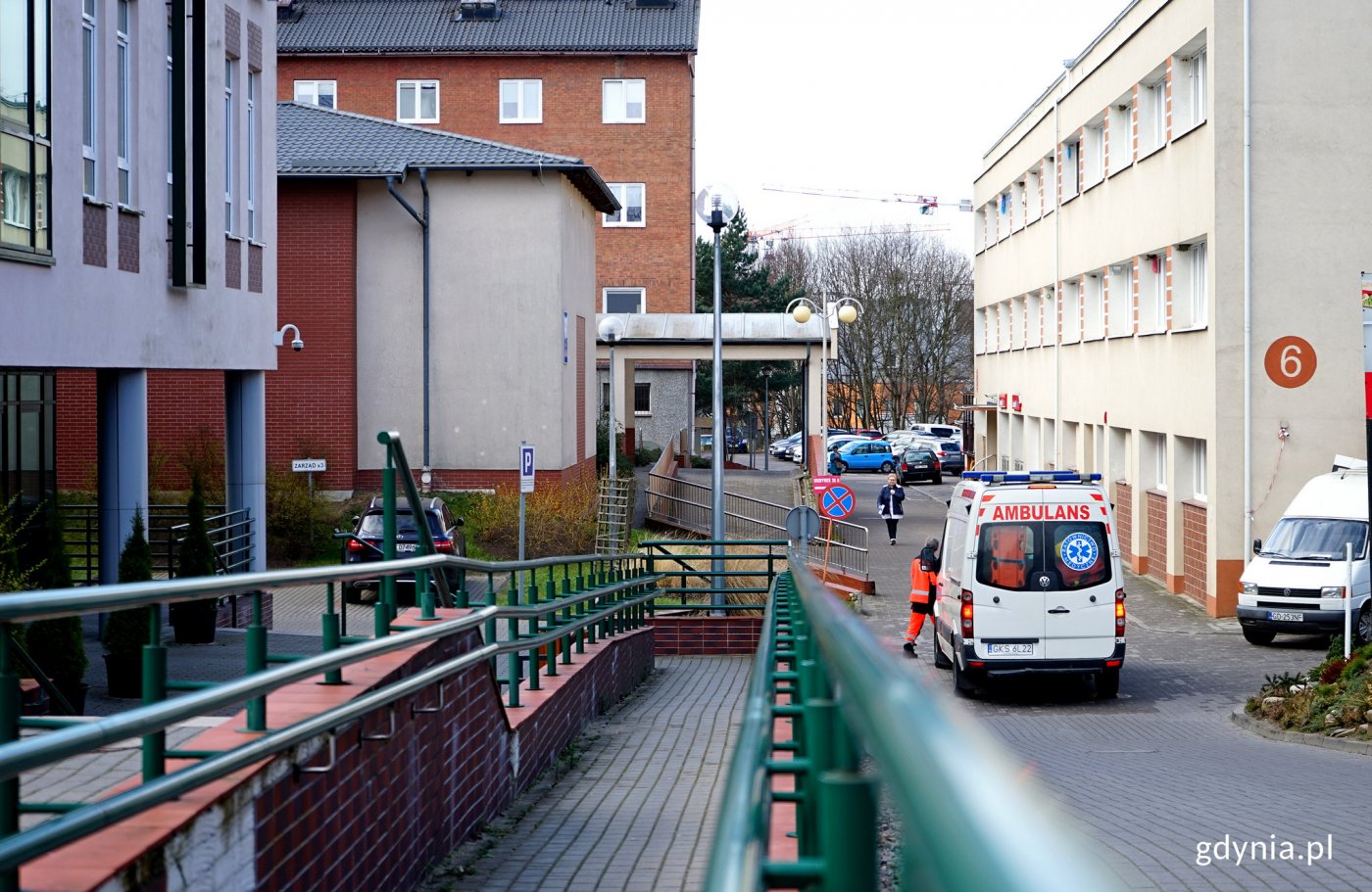 Budynki szpitala w Redłowie. Pośrodku droga wewnętrzna, stoi karetka pogotowia, idzie w jej kierunku ratownik medyczny