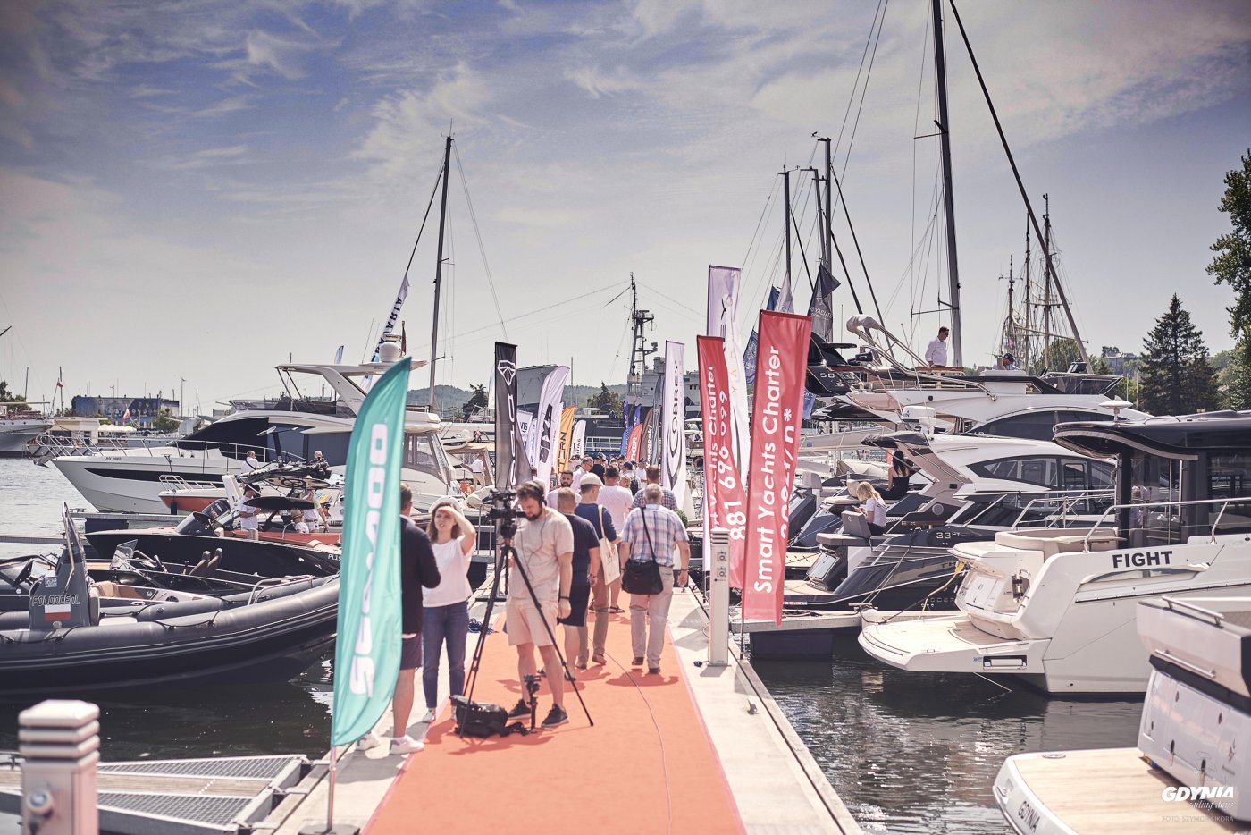 Keja w Marina Yacht Park w Gdyni. Ludzie spacerują po kei, zacumowane jachty w białym kolorze, słoneczny dzień