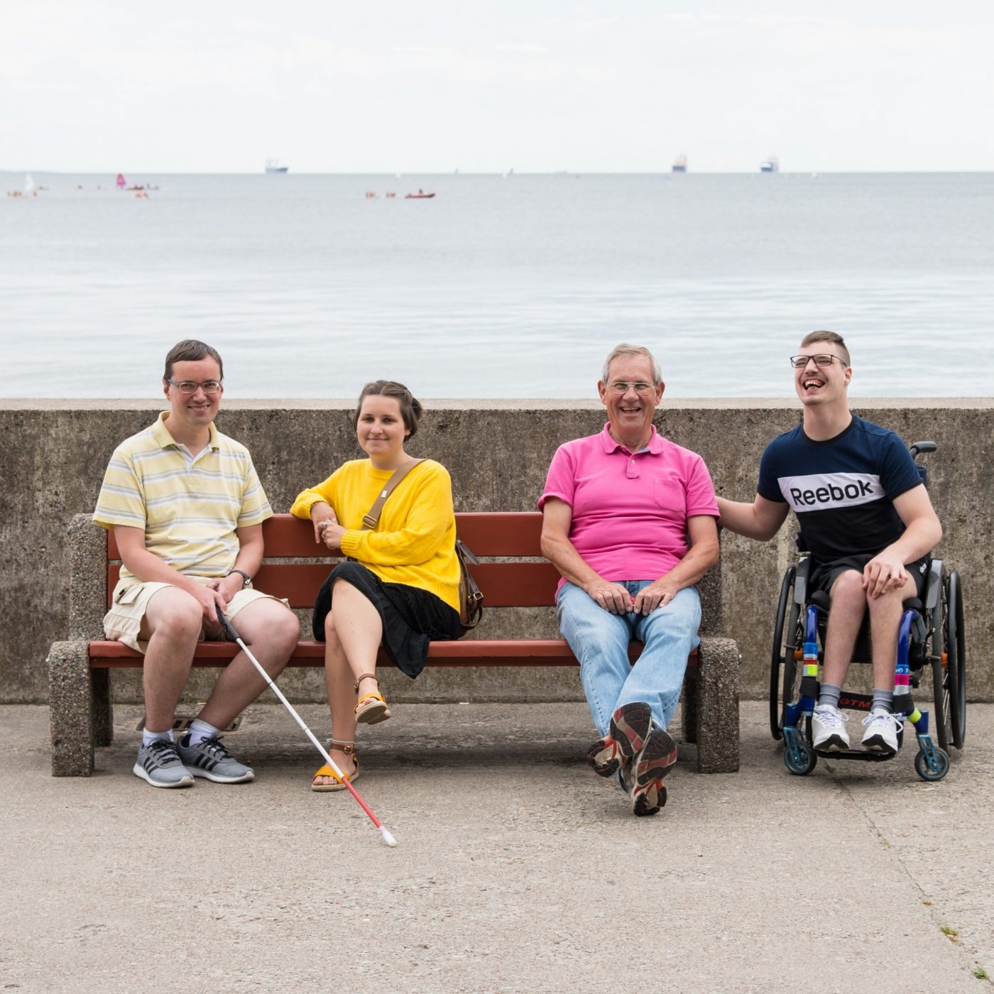 4 uśmiechnięte osoby siedzą na ławce na bulwarze.
W tym osoba poruszająca się na wózku,niewidoma i dwóch wolontariuszy.