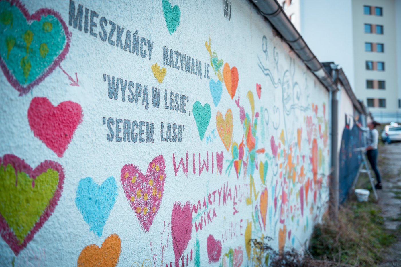 na zdjęciu fragment muralu na ul. Nauczycielskiej, otrzymany jest w jasnych kolorach, na pierwszym planie kolorowe serca i napis: mieszkańcy nazywają je: 'wyspą w lesie', 'sercem lasu'